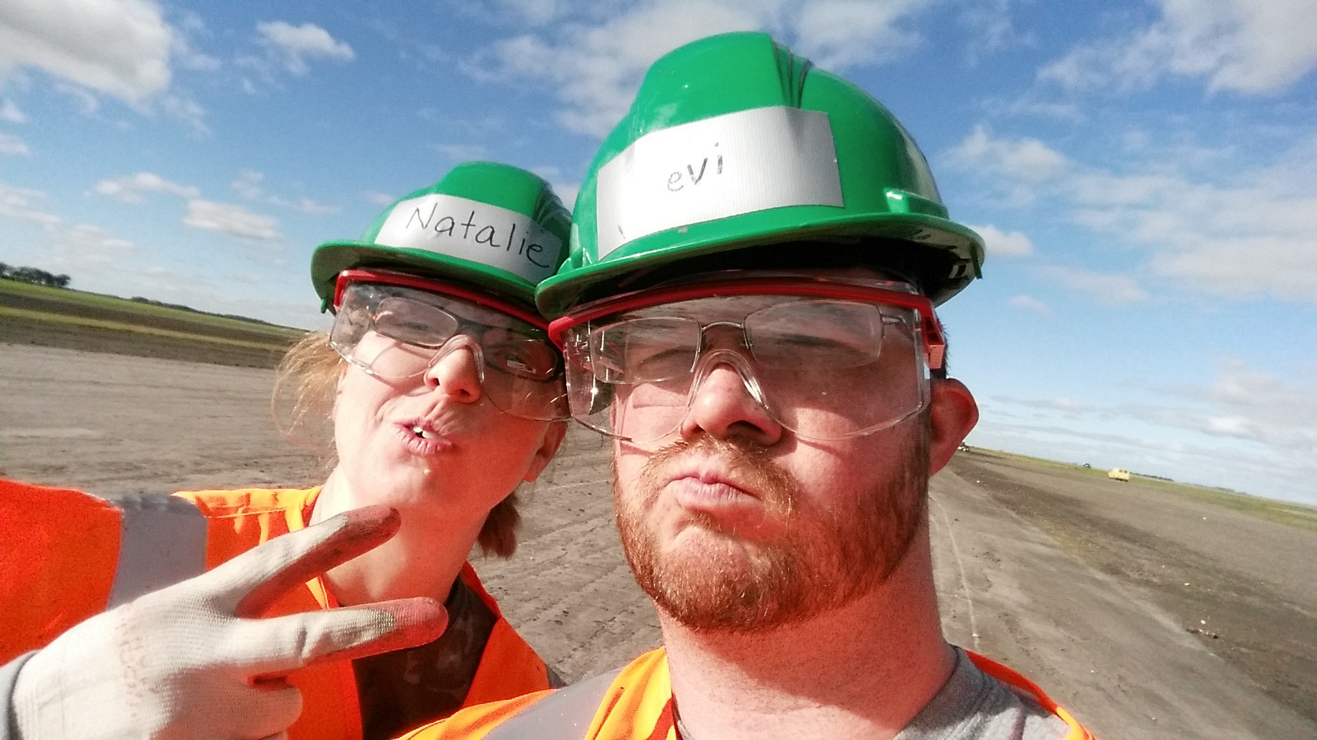 Workampers Levi and Natalie Henley taking a selfie wearing hard hats and safety vests at sugar beet harvest.