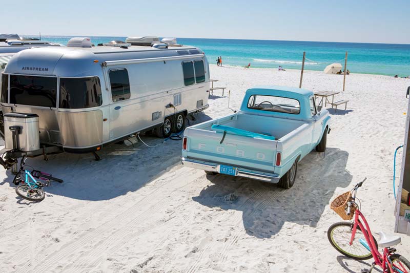 Airstream camper on white sand beach, next to an old ford pickup with a surfboard in the back, beautiful blue waters in the distance.