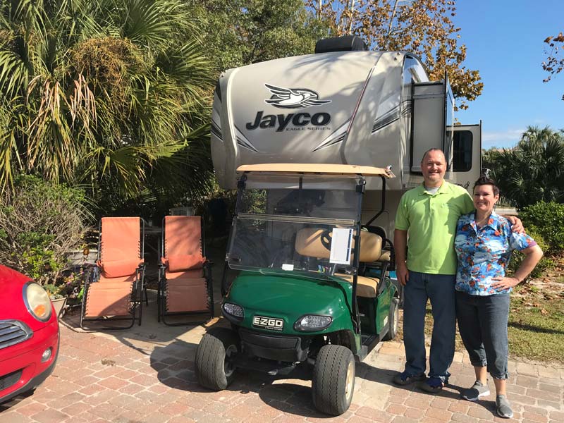 Workampers posing in front of Jayco 5th wheel at Camp Gulf RV site with golf cart to the side.