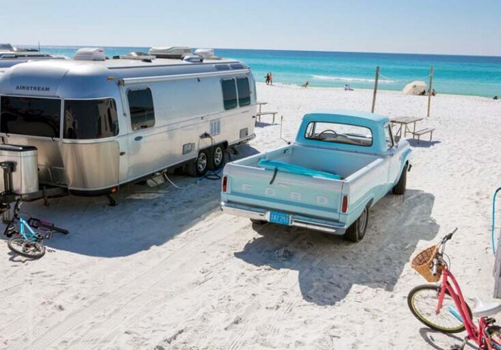 Airstream camper on white sand beach, next to an old ford pickup with a surfboard in the back, beautiful blue waters in the distance.