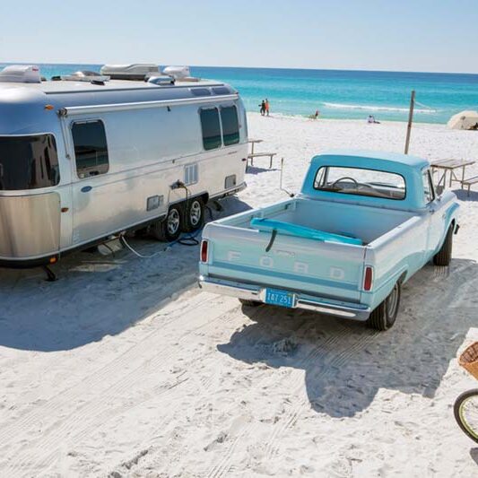 Airstream camper on white sand beach, next to an old ford pickup with a surfboard in the back, beautiful blue waters in the distance.