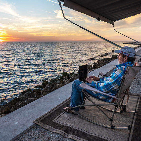 Man sitting with laptop in a chair outside a travel trailer next to an ocean