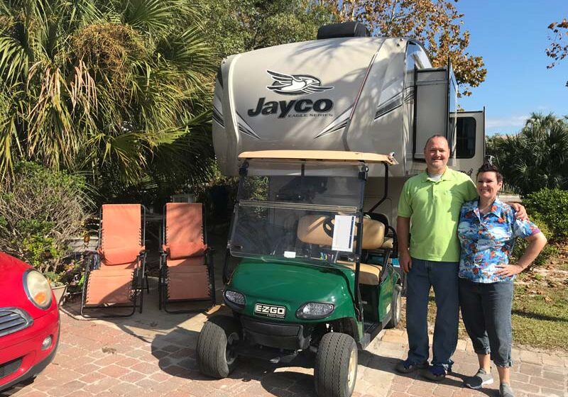 Workampers posing in front of Jayco 5th wheel at Camp Gulf RV site with golf cart to the side.