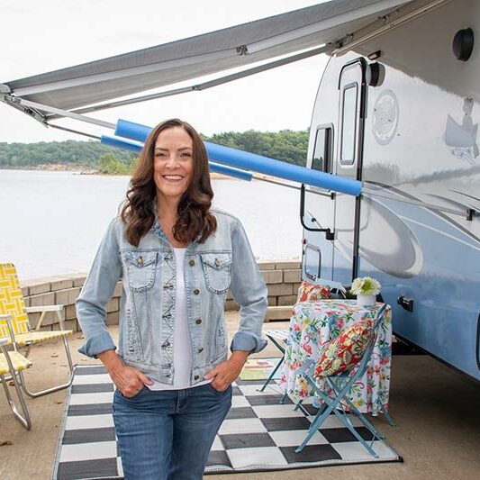 Workamper Brenda Pucket outside her RV at a lakeside location, illustrating her journey of turning her RV hobby into a career that empowers others.