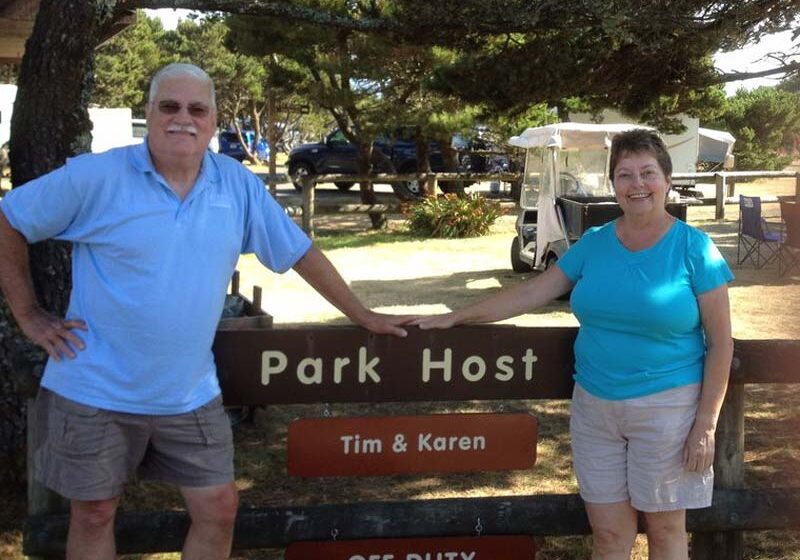 Workampers Tim and Karen Slack pose in front of park host sign in campground, golf cart in background.